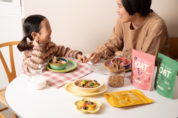 아침 식탁에서 그래놀라를 나눠 먹으며 웃는 부모와 아이, 옆에는 오리지널·딸기·바나나 맛 올댓오트 파우치가 놓여 있다